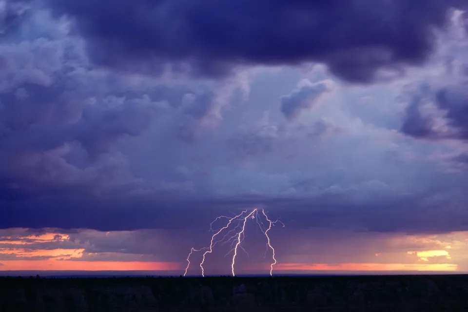 Multiple lightning bolts strike down from dark storm clouds over a silhouetted landscape at sunset.