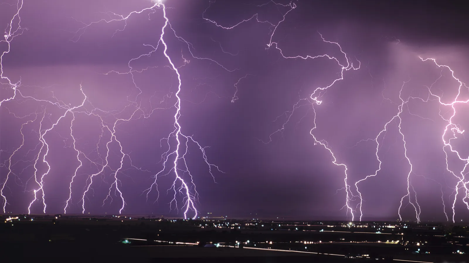 Multiple bright lightning bolts illuminate a dark, cloudy sky above a distant city.
