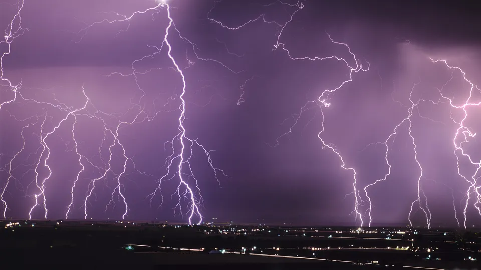 Multiple bright lightning bolts illuminate a dark, cloudy sky above a distant city.