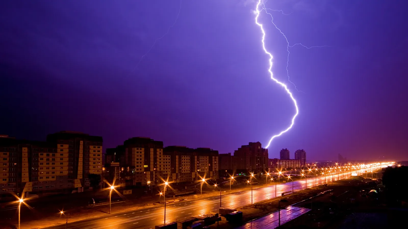 A bright lightning bolt strikes down from a dark purple sky onto a city skyline at night.