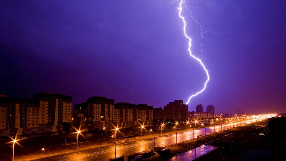 A bright lightning bolt strikes down from a dark purple sky onto a city skyline at night.