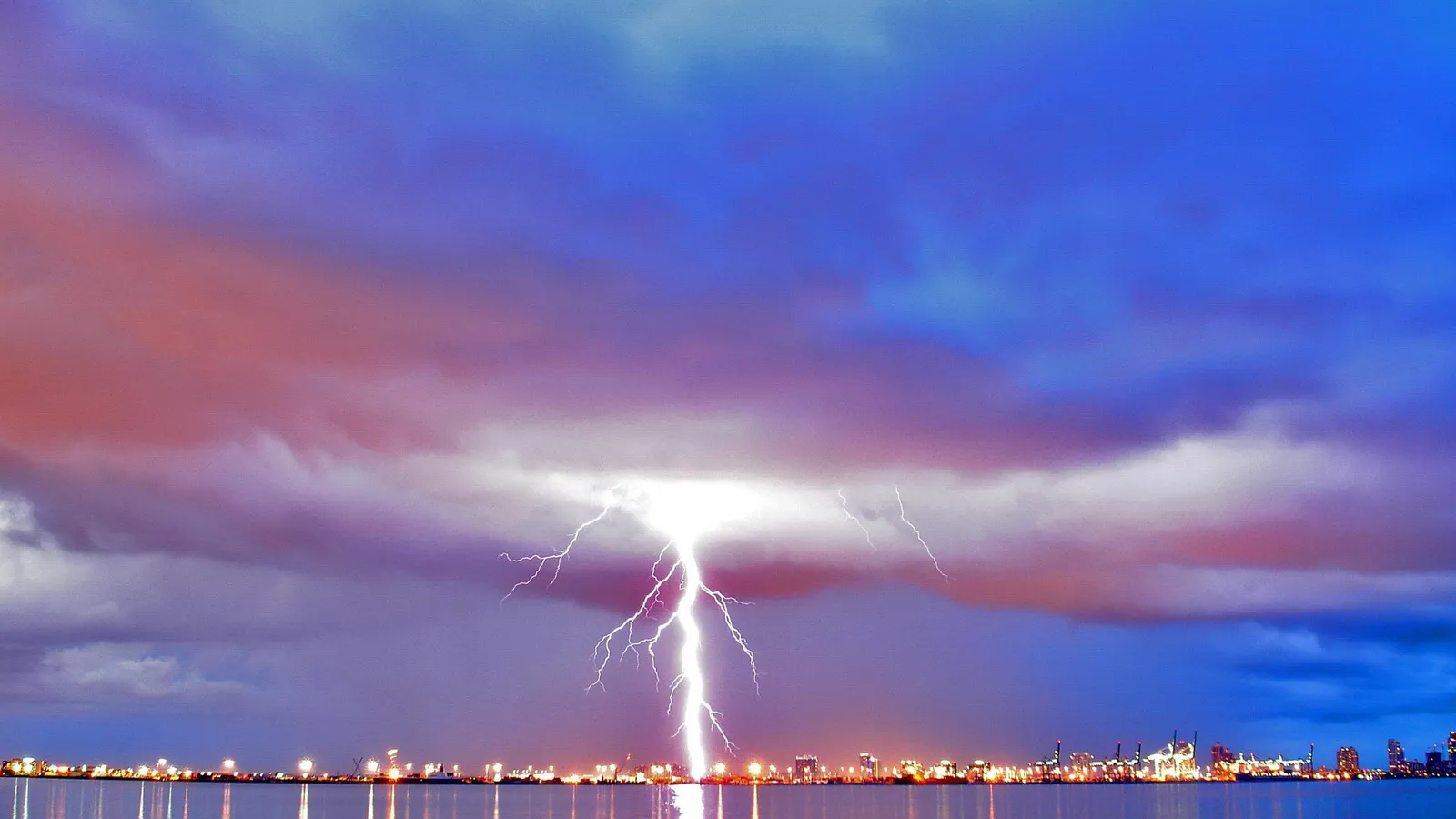 A bright lightning bolt strikes down from a stormy sky onto a city skyline at dusk.