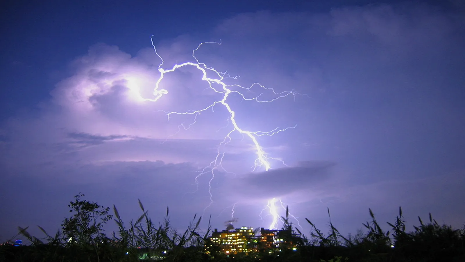 A bright lightning bolt flashes across a dark purple sky with clouds, illuminating a distant city.