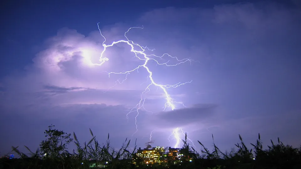 A bright lightning bolt flashes across a dark purple sky with clouds, illuminating a distant city.