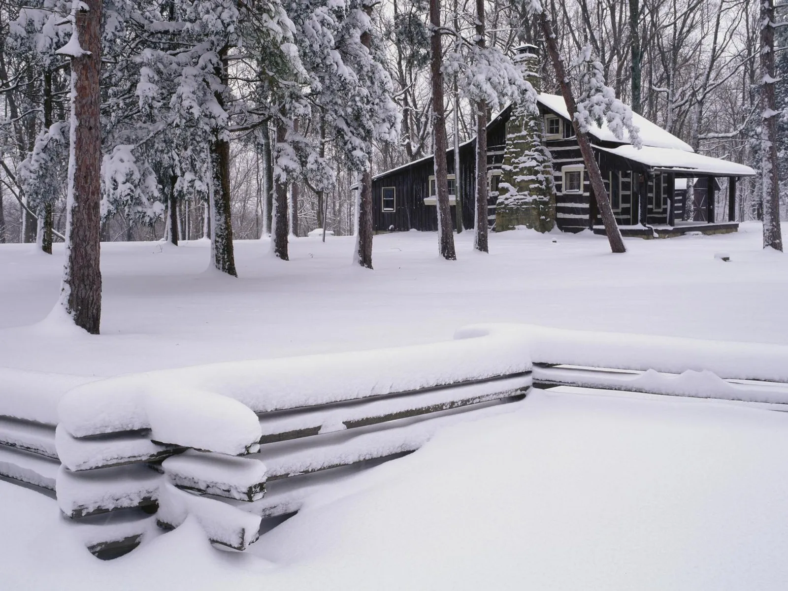 A dark log cabin is nestled in a snowy forest with a split rail fence in the foreground.
