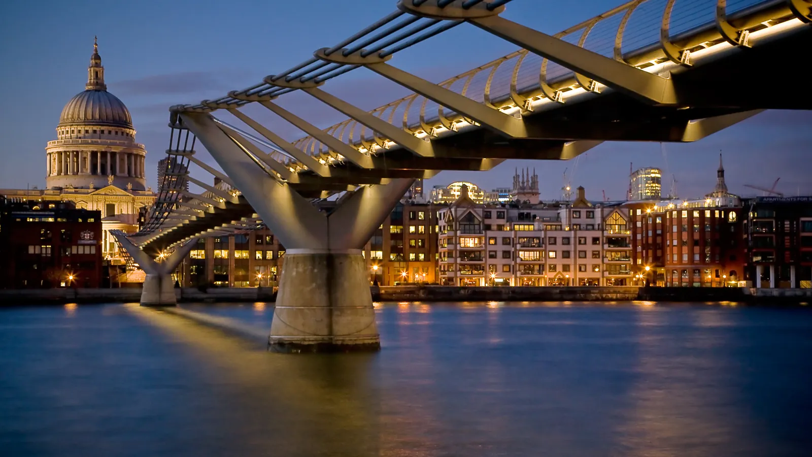 The Millennium Bridge spans the River Thames in London, with St. Paul's Cathedral and city buildings in the background at dusk.