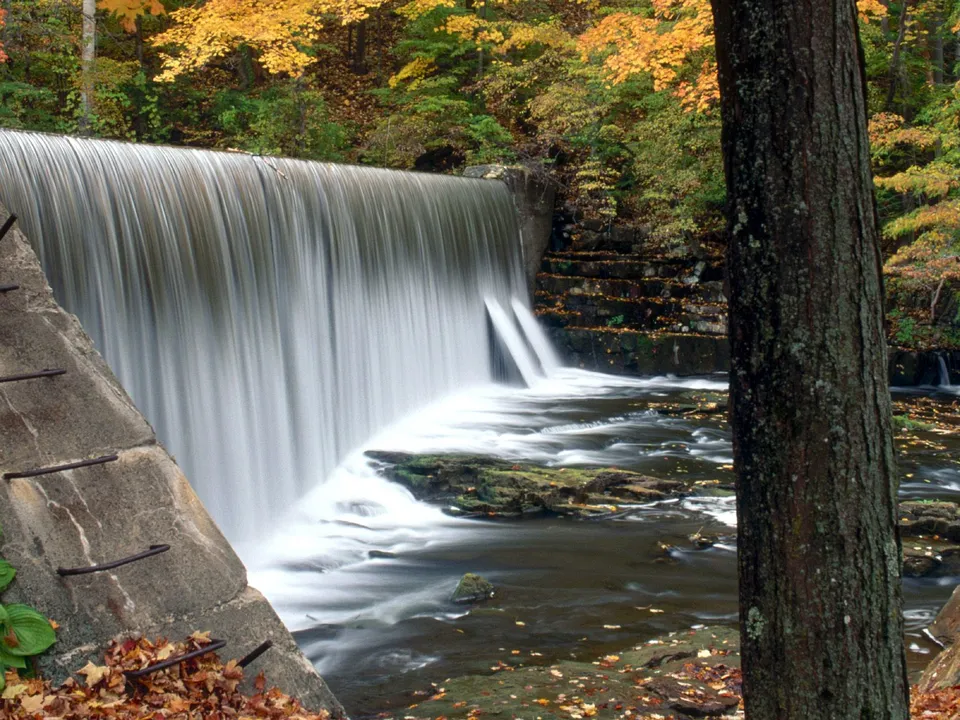 A wide dam waterfall with a silky effect in autumn, surrounded by colorful trees.