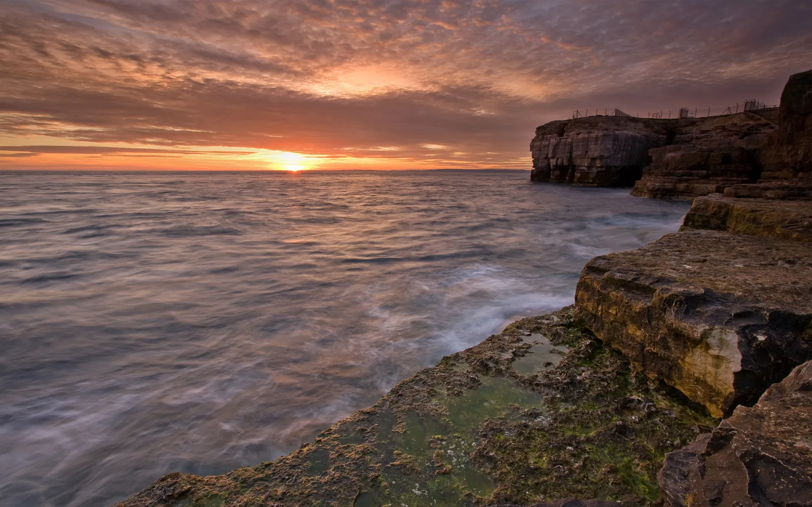 A long exposure shot of the sun setting over layered coastal cliffs and the ocean.