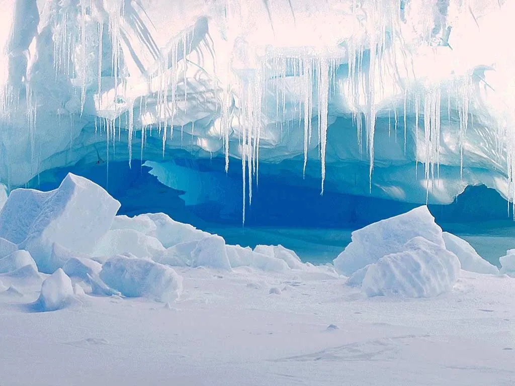 Long icicles hang from the ceiling of a blue ice cave with large ice chunks in the foreground.