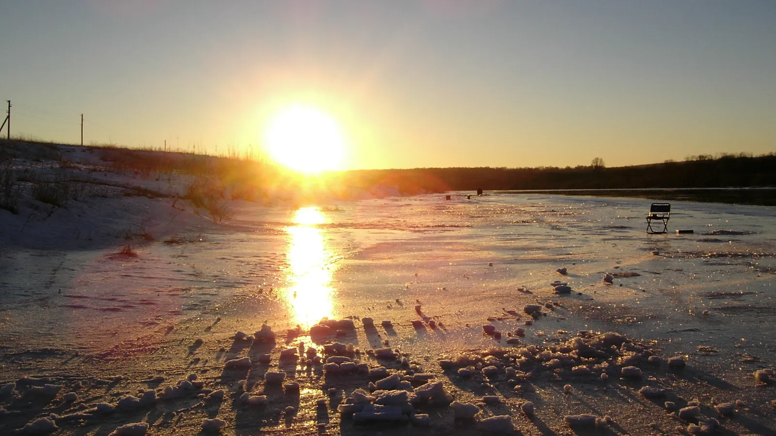 A bright winter sun low on the horizon casts a golden reflection on a frozen lake surface.
