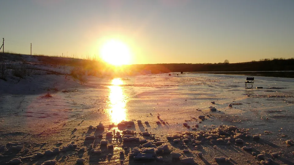 A bright winter sun low on the horizon casts a golden reflection on a frozen lake surface.