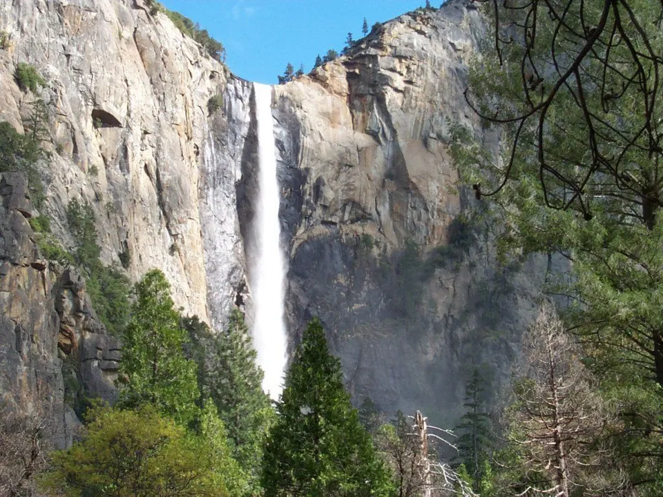 A tall waterfall descends a vast, rugged cliff, framed by lush green trees on either side.