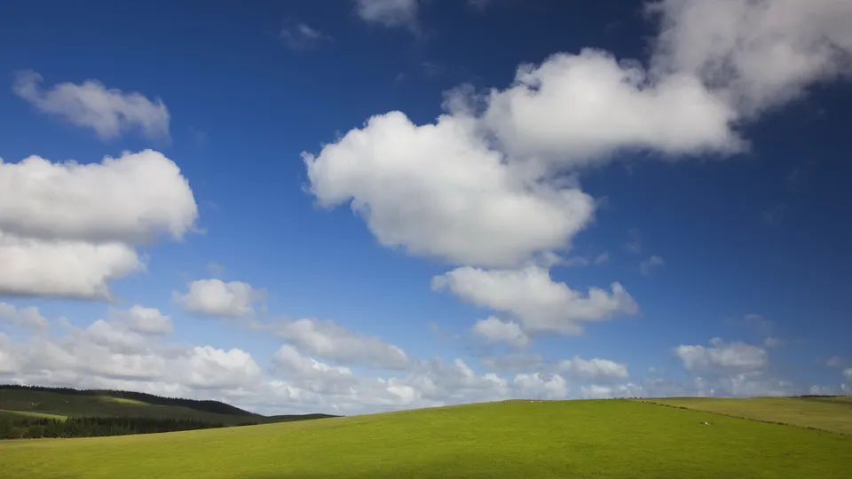 Lush green fields stretch across rolling hills under a vibrant blue sky dotted with white cumulus clouds.