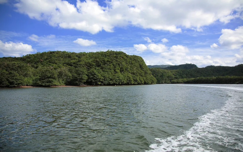 A wide river flows past lush green hills under a blue sky with white clouds.