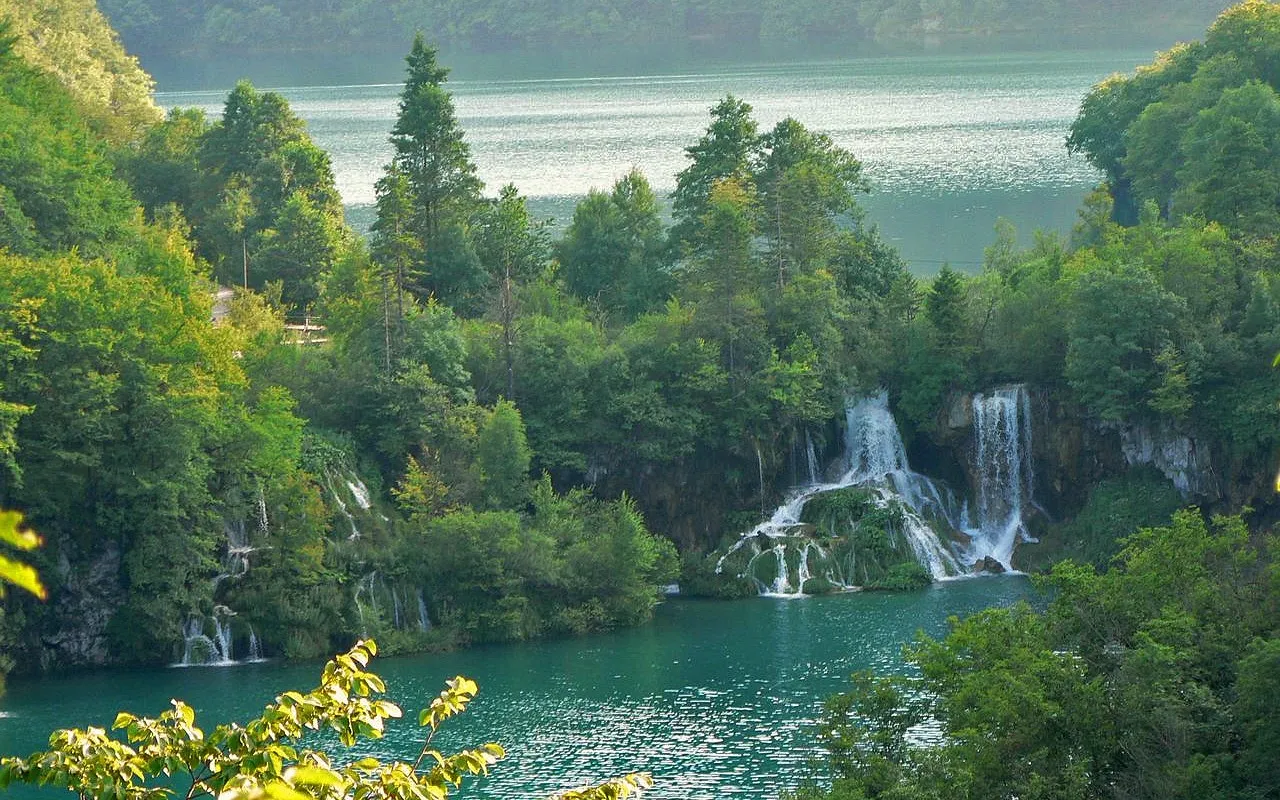 A series of waterfalls cascade down mossy rocks into a clear turquoise lake, surrounded by dense green forest.