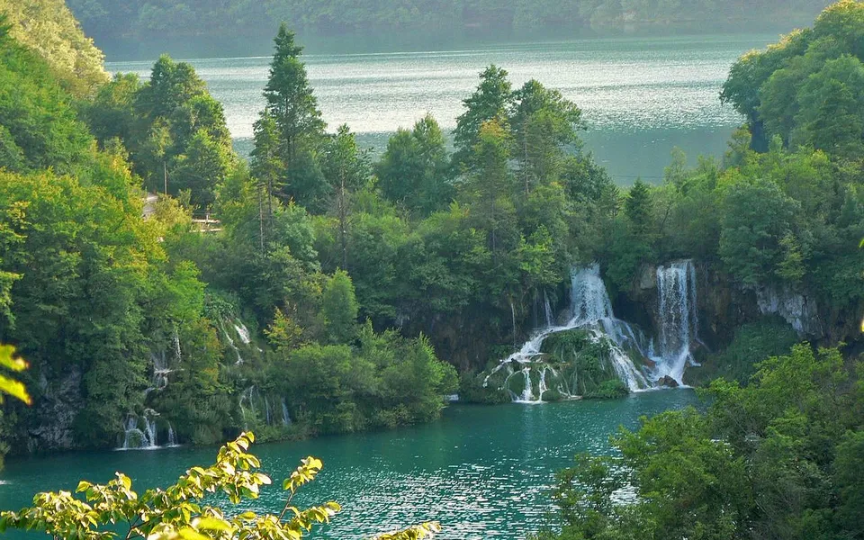 A series of waterfalls cascade down mossy rocks into a clear turquoise lake, surrounded by dense green forest.