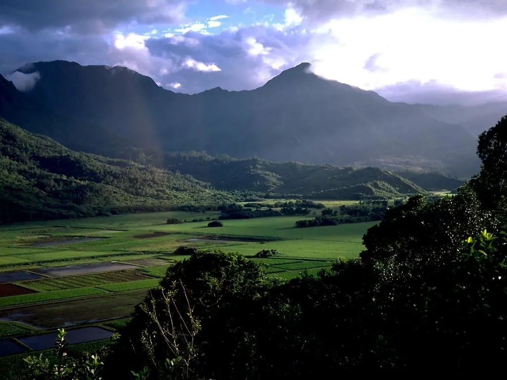 A wide valley with green fields and mountains under a cloudy sky with sunbeams.