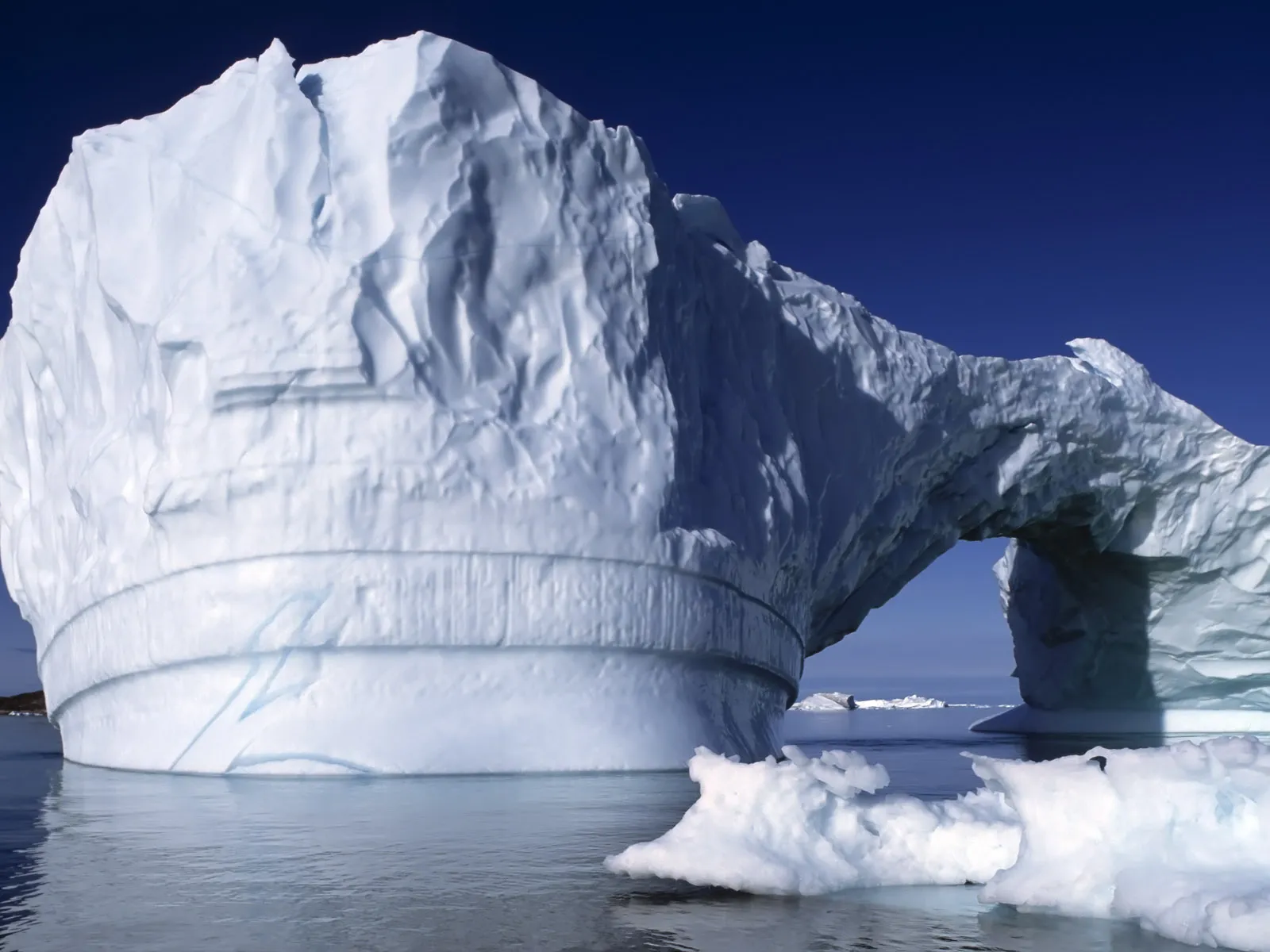 An iceberg with a prominent arch is illuminated by bright daylight, with smaller ice formations in the foreground.