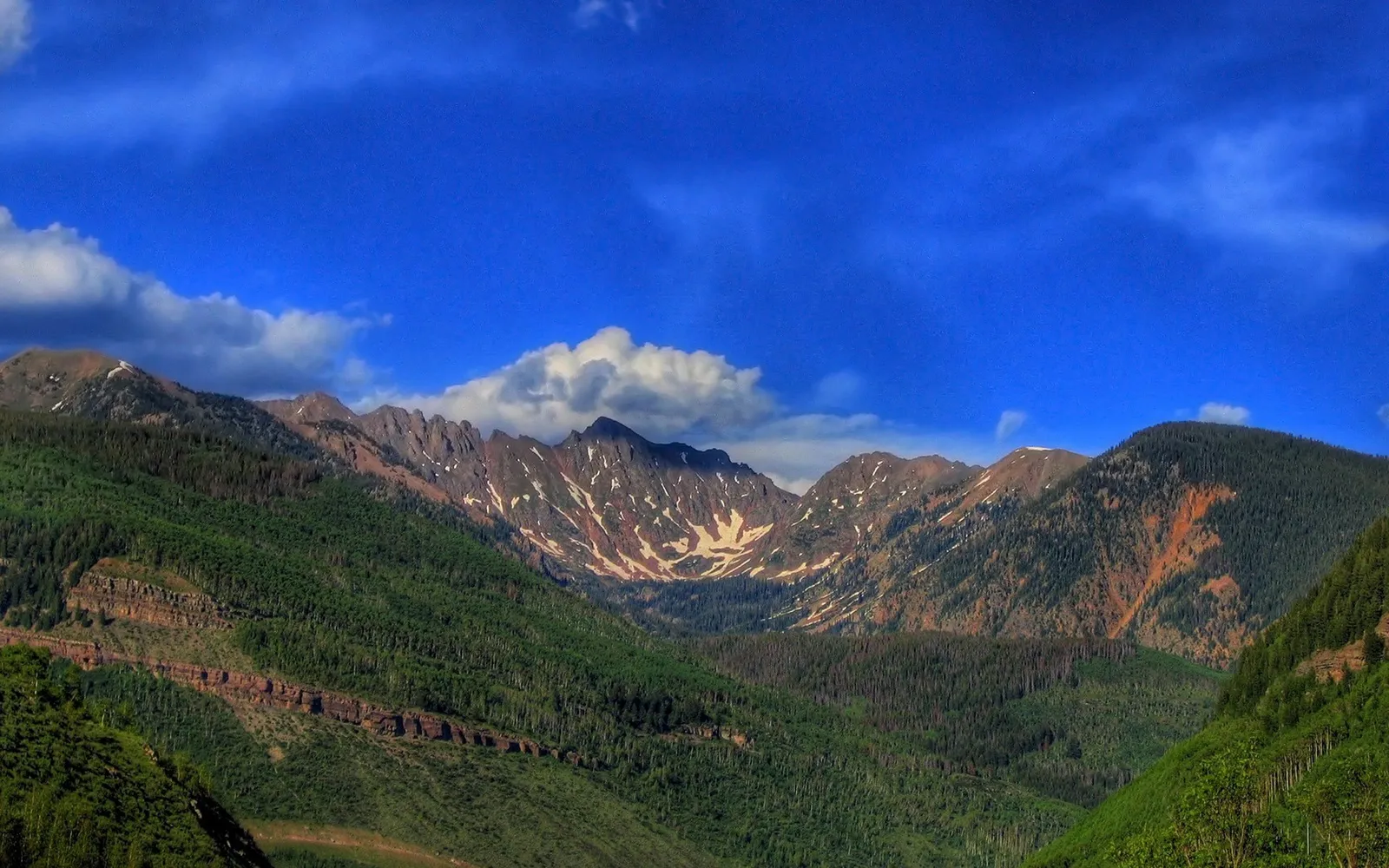 A panoramic view of rugged, tree-covered mountains under a bright blue sky with scattered clouds.