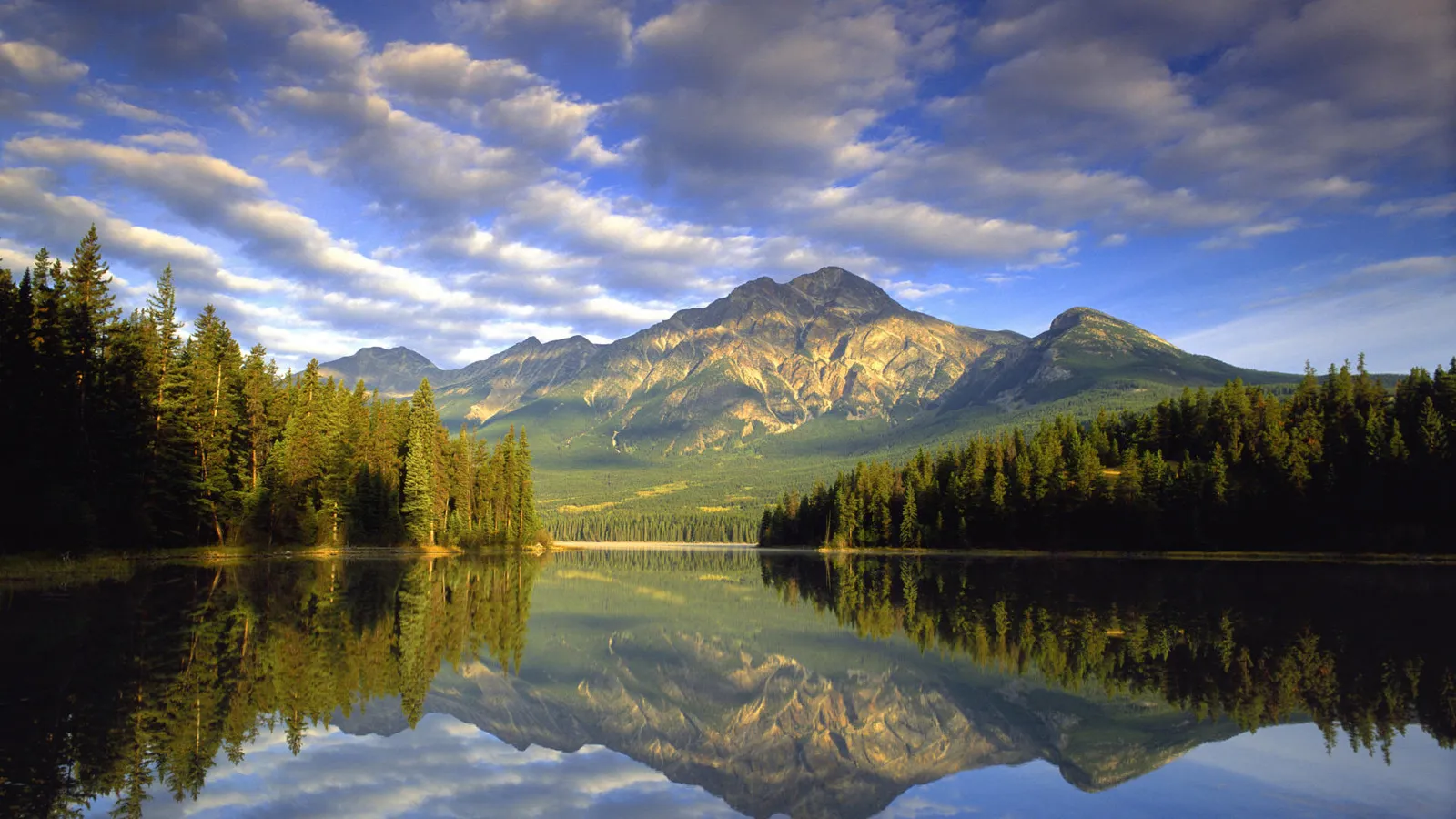 A majestic mountain is reflected in a calm lake surrounded by pine forests under a cloudy sky.