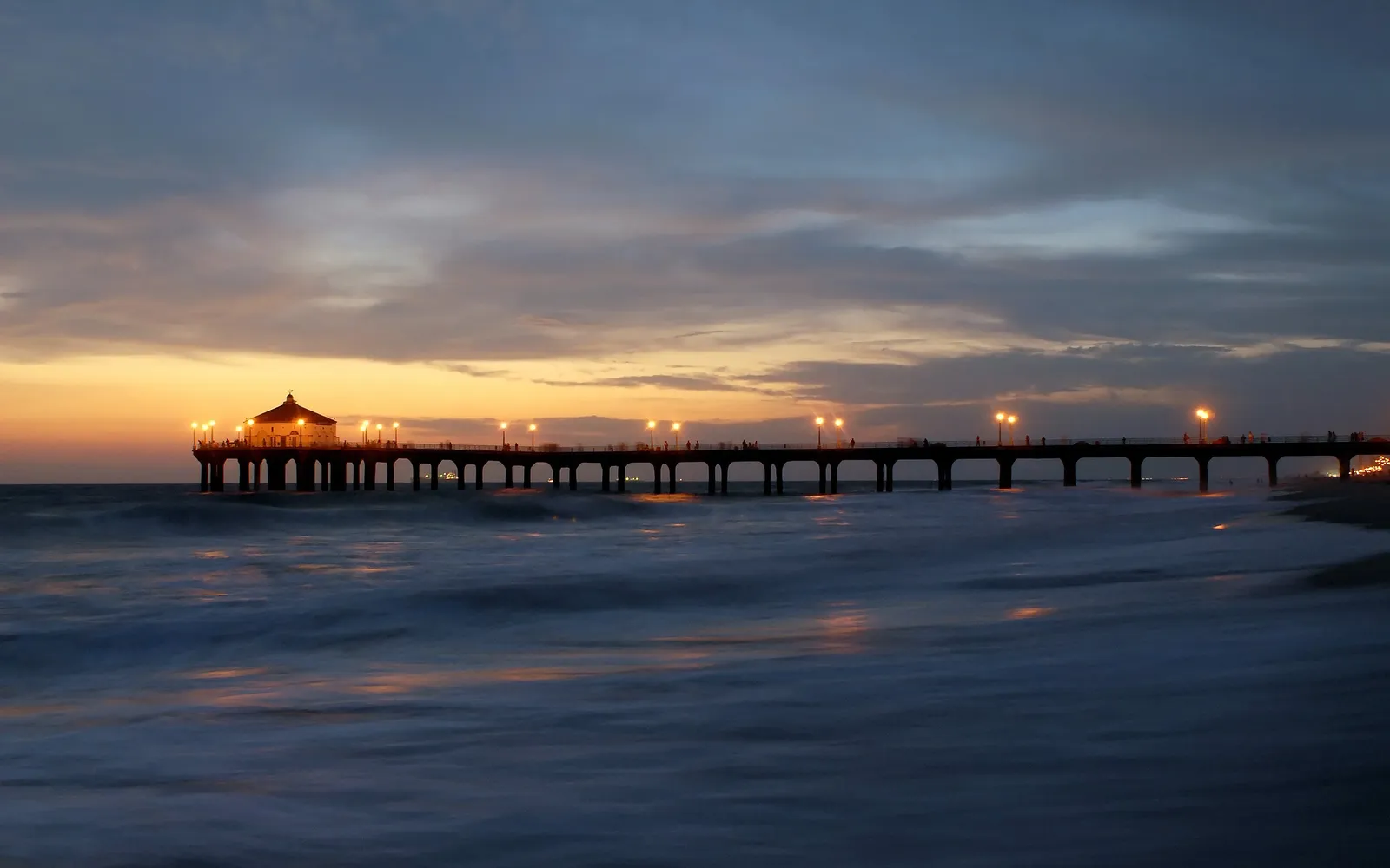 A long pier with lights extends over the ocean at sunset, with waves crashing on the shore.