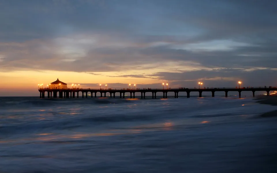 A long pier with lights extends over the ocean at sunset, with waves crashing on the shore.