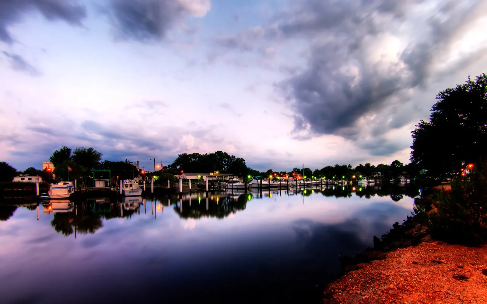 Boats are docked in a marina on a calm body of water reflecting the twilight sky and lights.