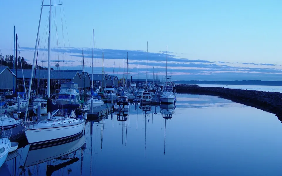 Sailboats and motorboats docked in a marina at dusk, with their reflections visible in the calm water.