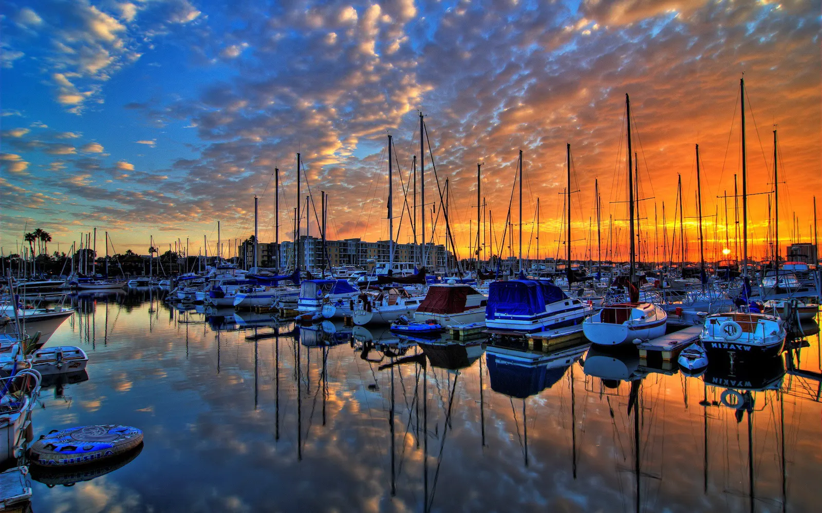 A marina filled with sailboats is reflected in the calm water during a vibrant sunset with dramatic clouds.