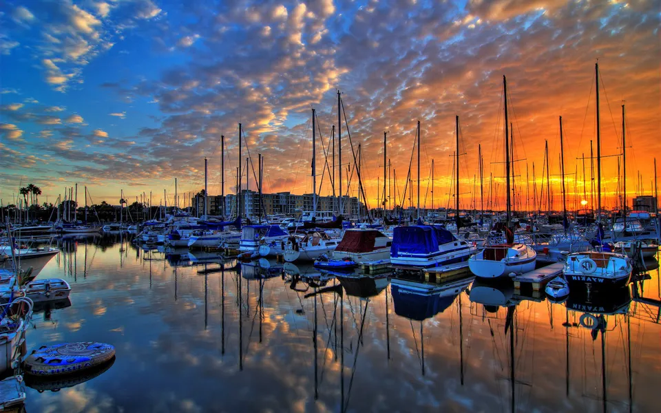 A marina filled with sailboats is reflected in the calm water during a vibrant sunset with dramatic clouds.