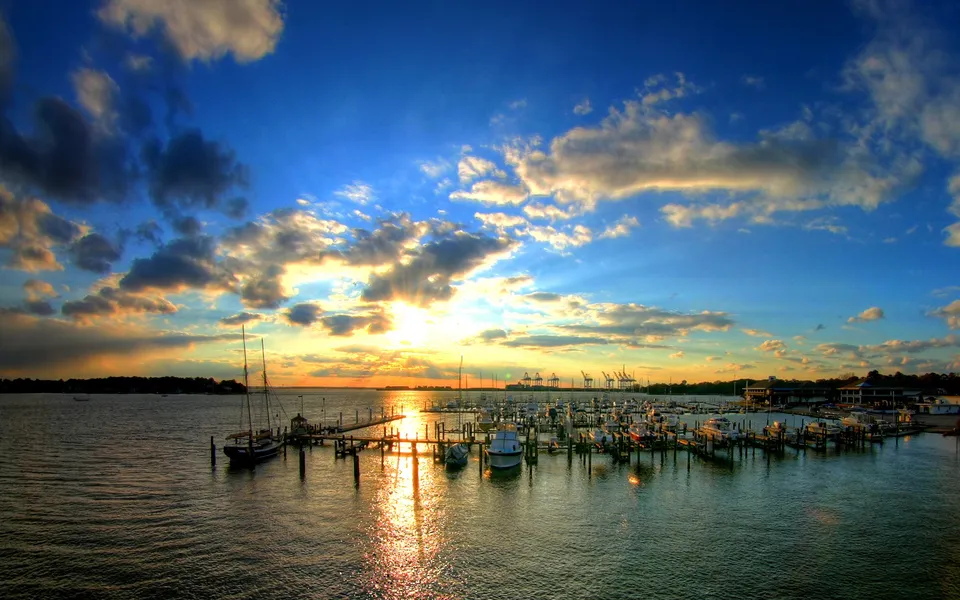 A vibrant sunset with golden sun rays breaking through dramatic clouds over a marina filled with boats.