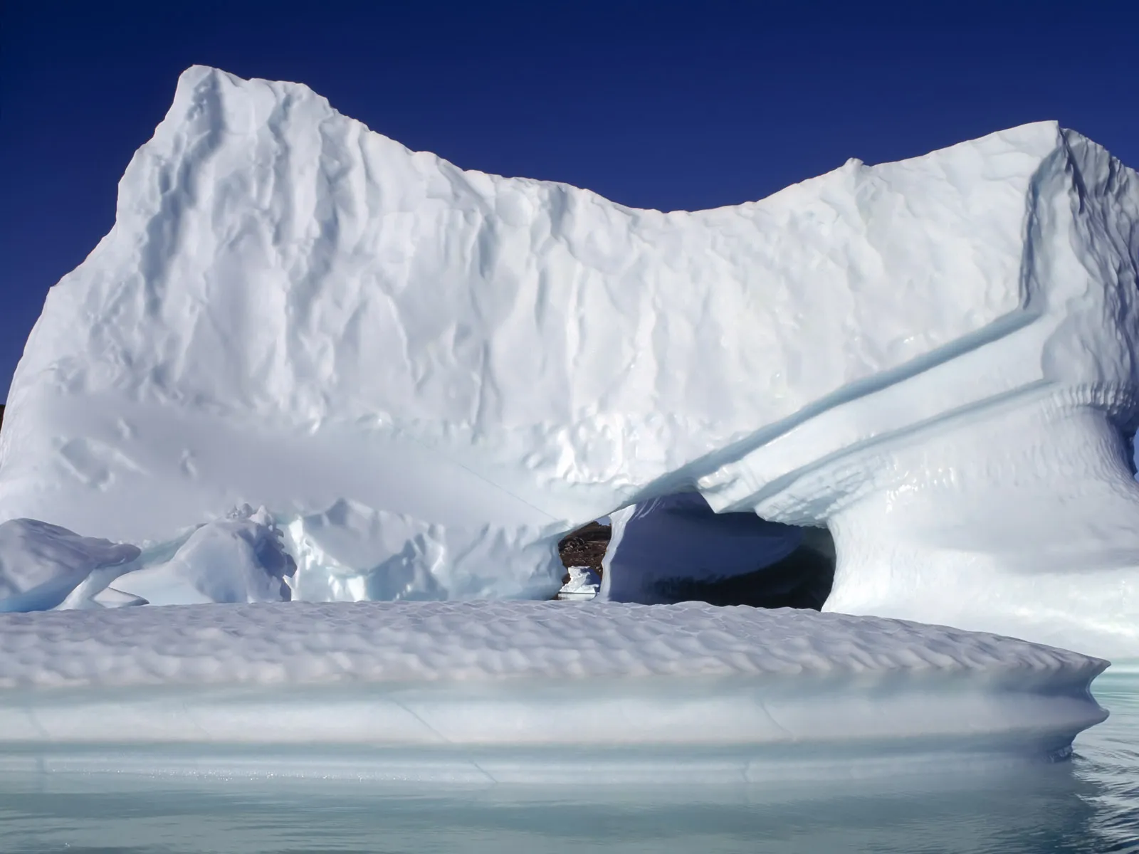 A large iceberg with a natural archway is set against a deep blue sky, with calm water in the foreground.