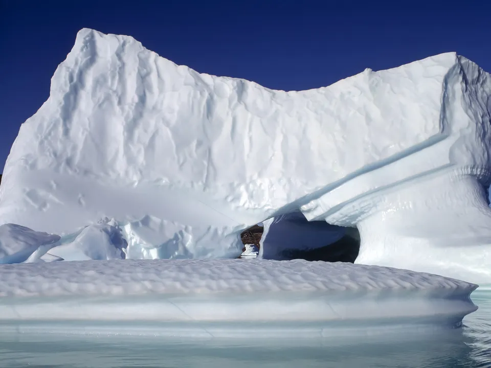 A large iceberg with a natural archway is set against a deep blue sky, with calm water in the foreground.