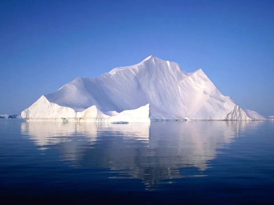 A large, white iceberg is reflected perfectly in the calm, deep blue water of the Arctic Ocean under a clear blue sky.