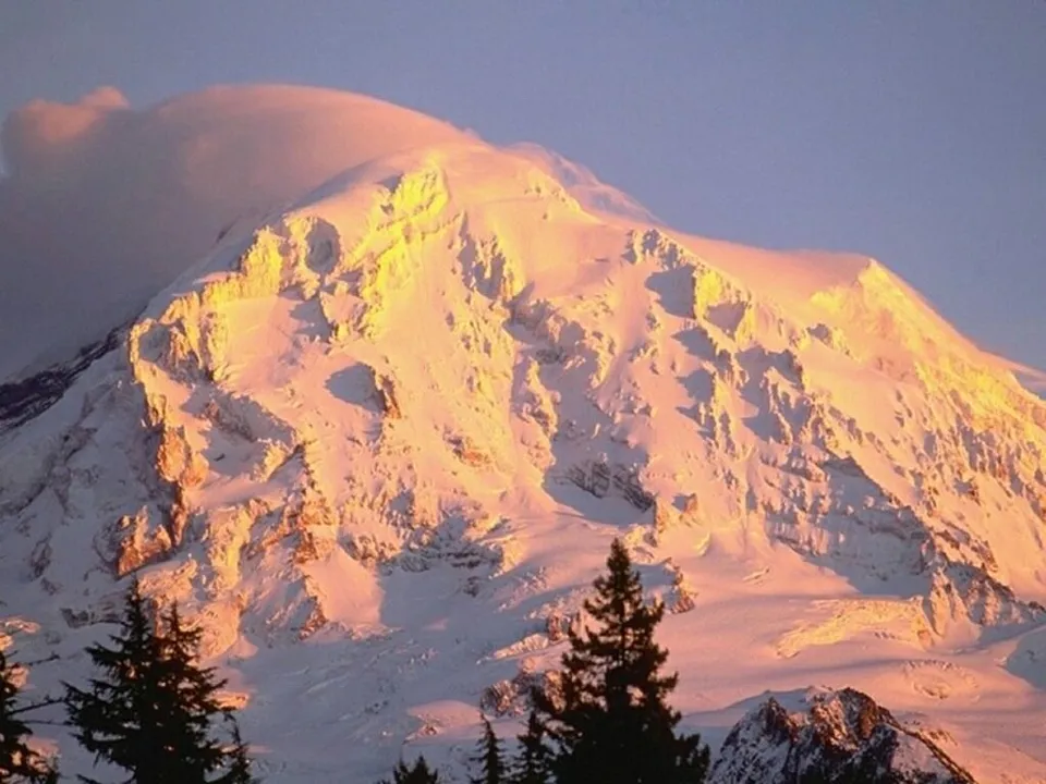 A massive, snow-covered mountain is bathed in the warm, golden light of sunrise, highlighting its rugged terrain.