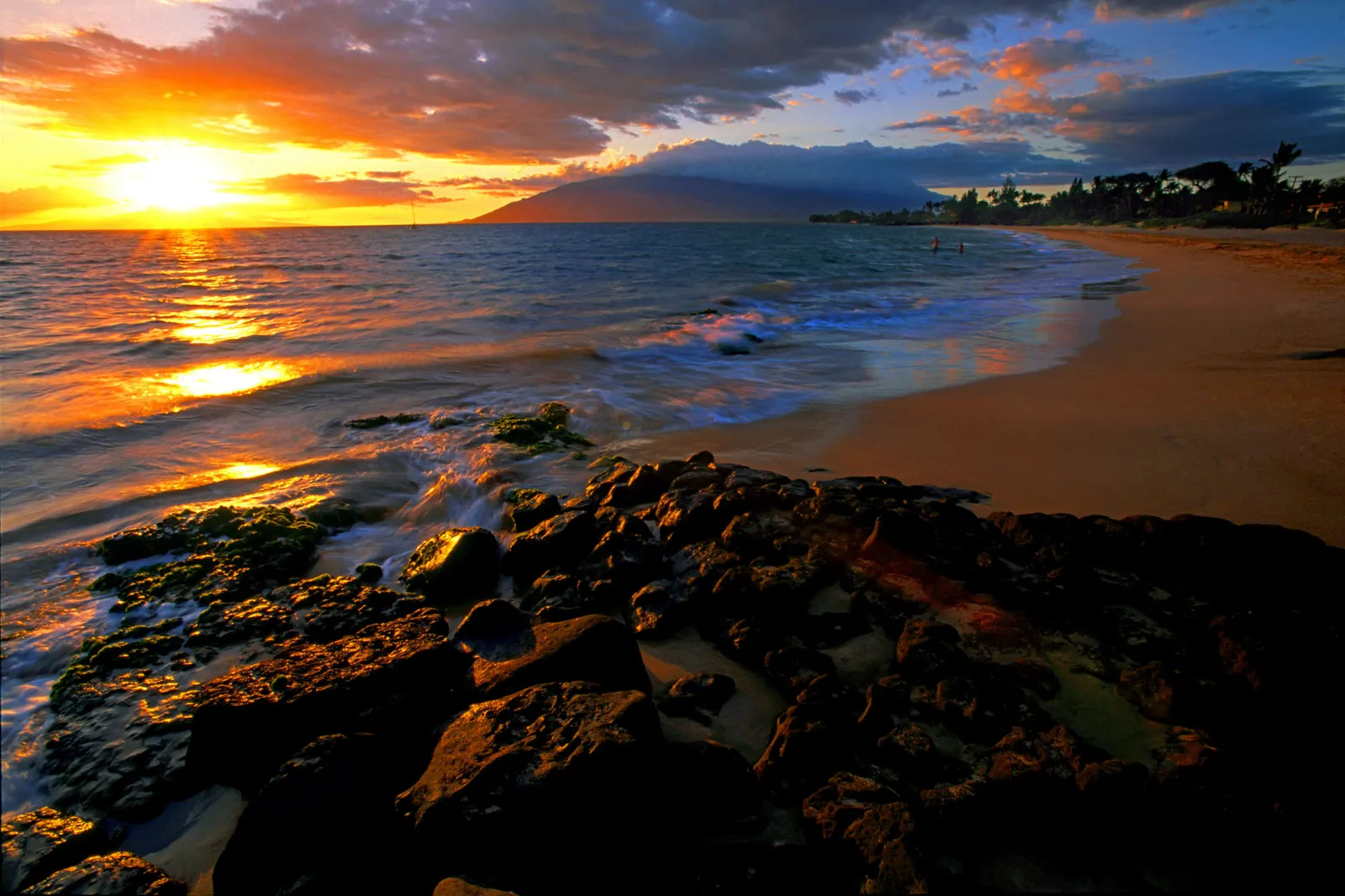A vibrant sunset casts golden light across the ocean and a sandy beach with rocky foreground.