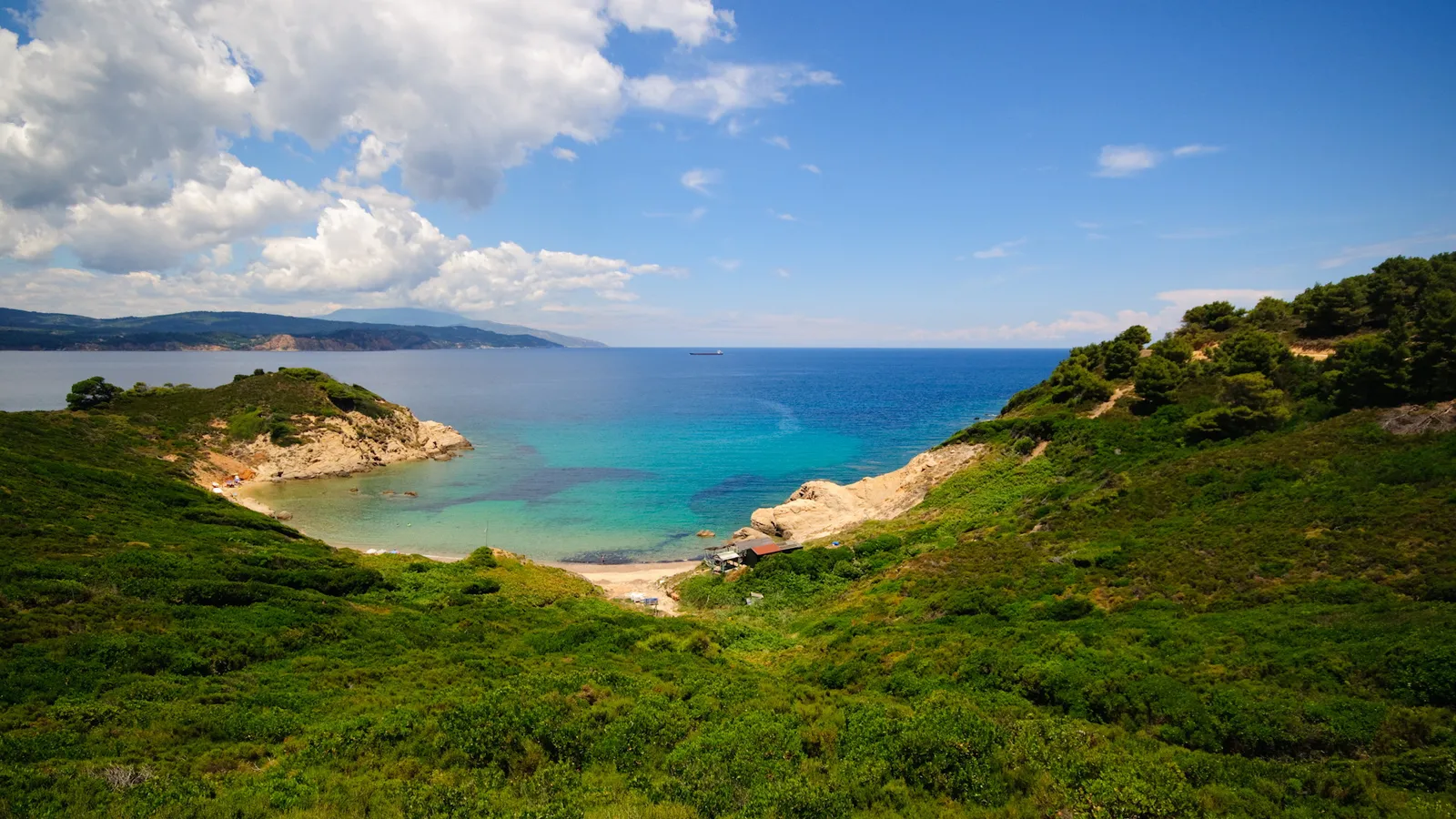 A view of a secluded cove with clear turquoise water, sandy beach, and lush green hills under a blue sky with white clouds.