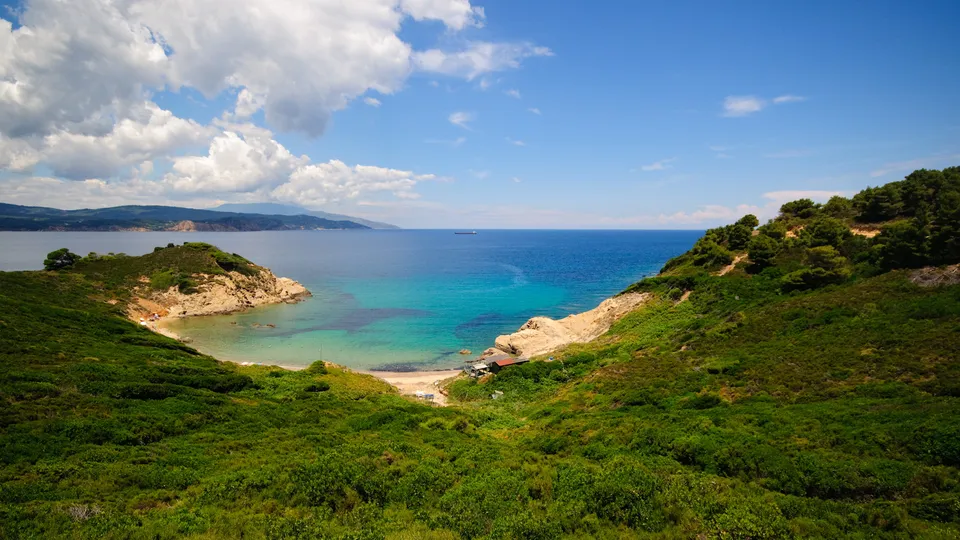 A view of a secluded cove with clear turquoise water, sandy beach, and lush green hills under a blue sky with white clouds.