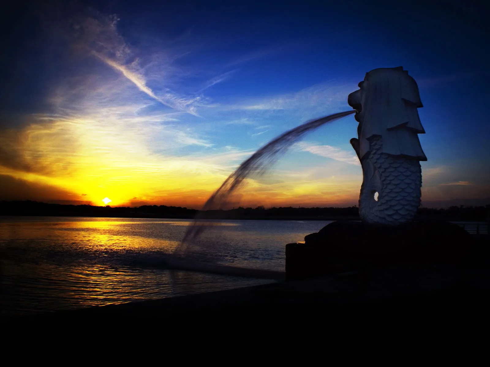 The Merlion statue in Singapore spouts water into the sea at sunset.