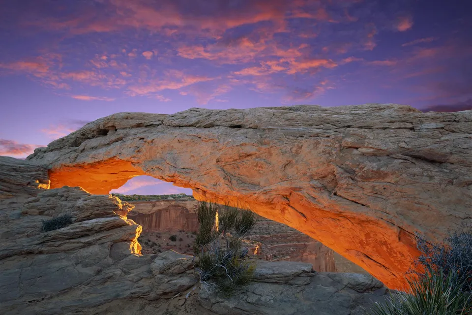 Mesa Arch in Canyonlands National Park glows orange from sunrise, with a purple and pink sky above.