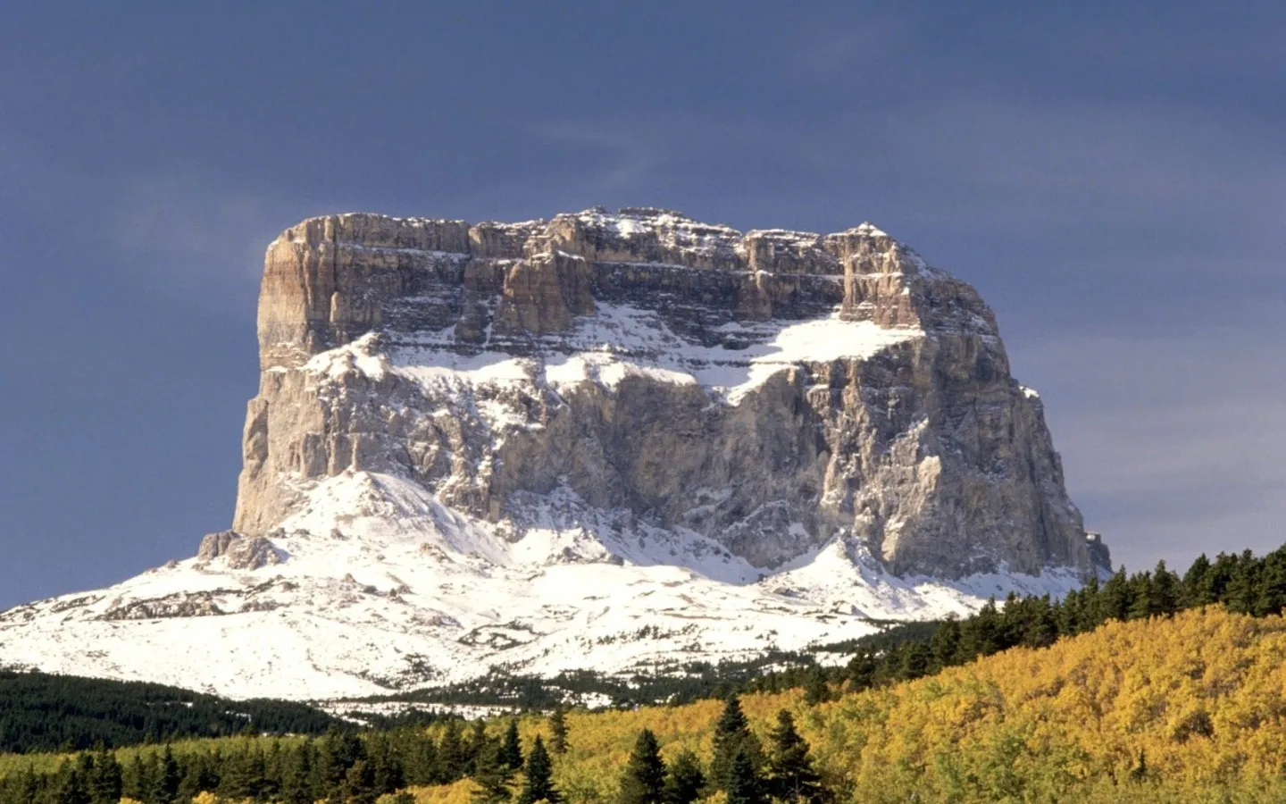 A large, flat-topped mountain with snow-covered slopes and a forest in the foreground.