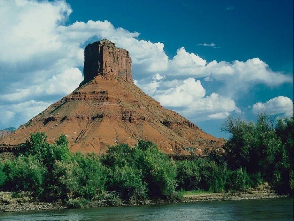 A large, flat-topped mesa with reddish-brown rock layers rises above a green riverbank and a wide river.