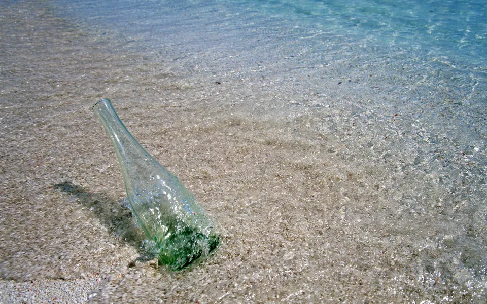 A glass bottle lies on its side in shallow, clear ocean water on a sandy beach.