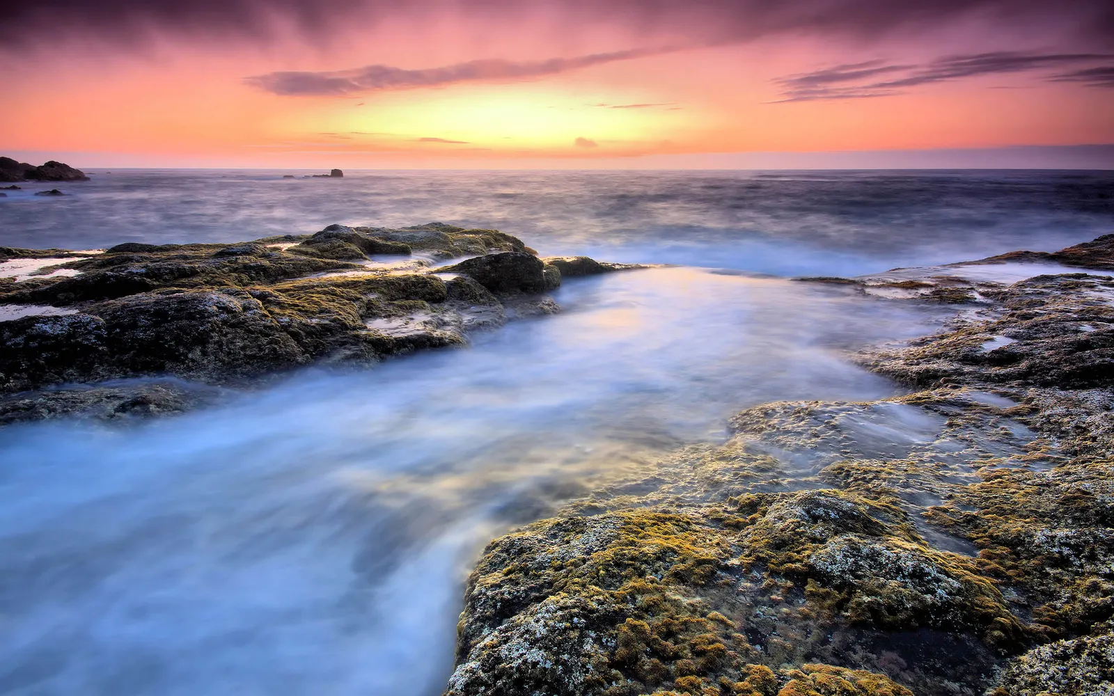 Smooth, milky water flows around dark, moss-covered rocks during a pink and orange sunset.