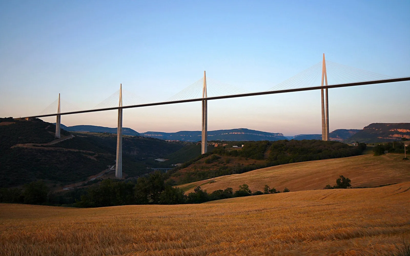 A long cable-stayed bridge stretches across a valley with rolling hills and a field of wheat in the foreground.
