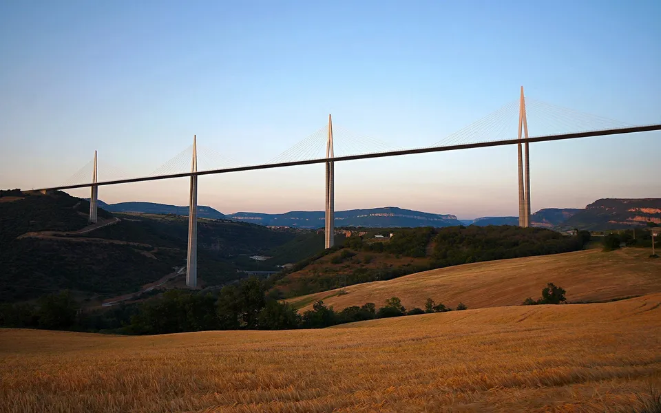 A long cable-stayed bridge stretches across a valley with rolling hills and a field of wheat in the foreground.