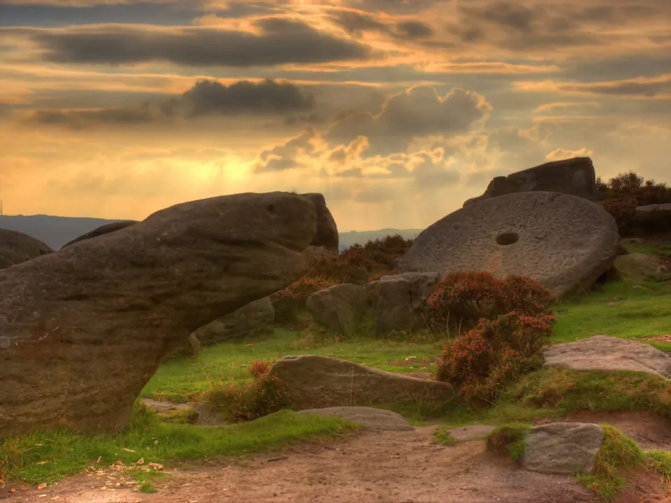 A large, circular stone with a hole in the center rests on a grassy hillside with heather and other rocks.