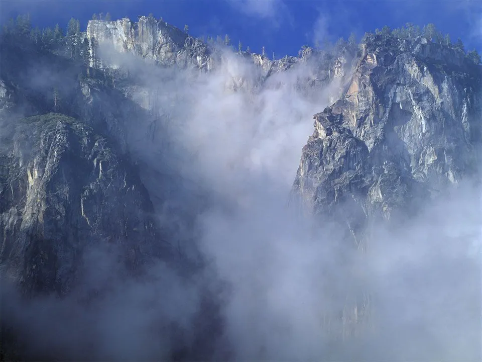 Towering granite cliffs are partially obscured by swirling mist and clouds against a bright blue sky.