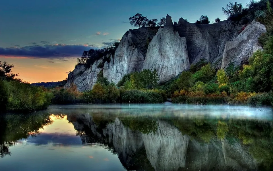 A serene lake reflects dramatic, eroded cliffs under a colorful dawn sky with mist rising from the water.