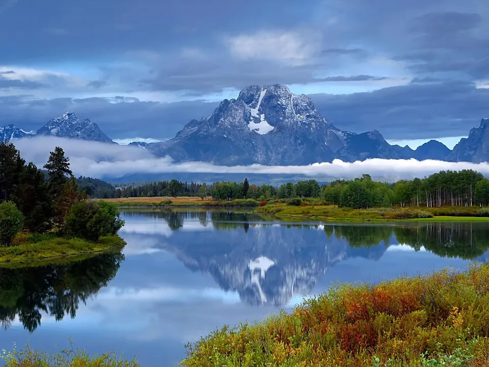 Misty clouds surround snow-capped mountain peaks reflected in a wide river.